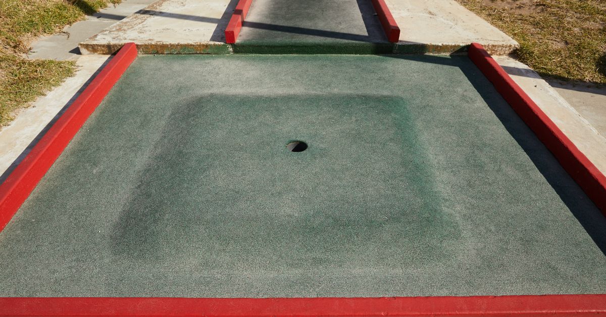 A red-bordered artificial-turf mini golf hole with a single cup, seen from above in bright sunlight.