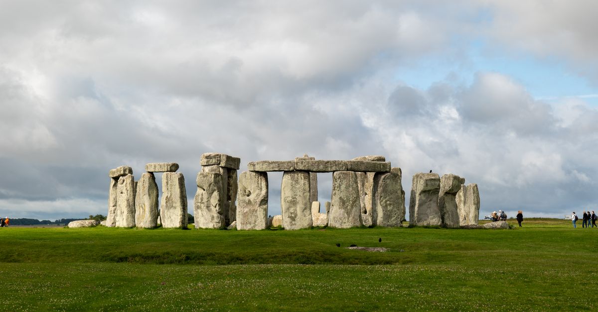 Stonehenge's stone circle with trilithons and lintels under a cloudy sky, visitors in the distance.