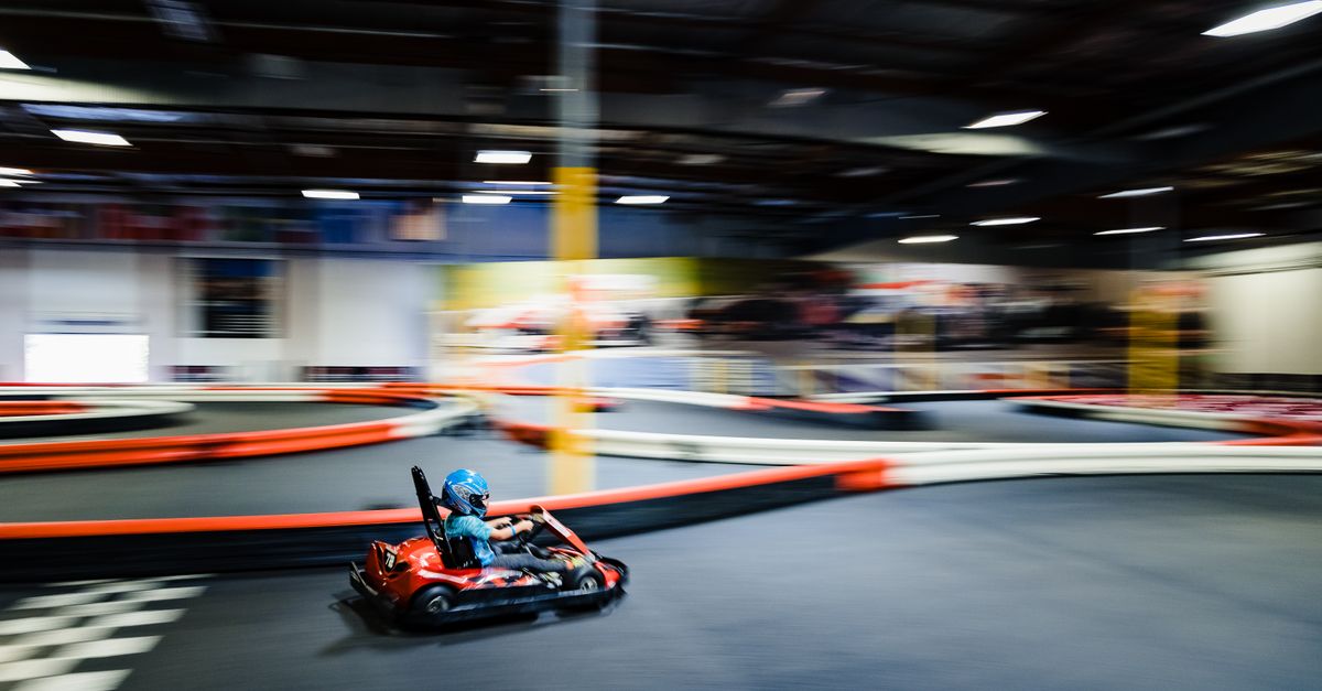 A helmeted go-kart driver taking a corner at speed on an indoor track, motion-blurred.