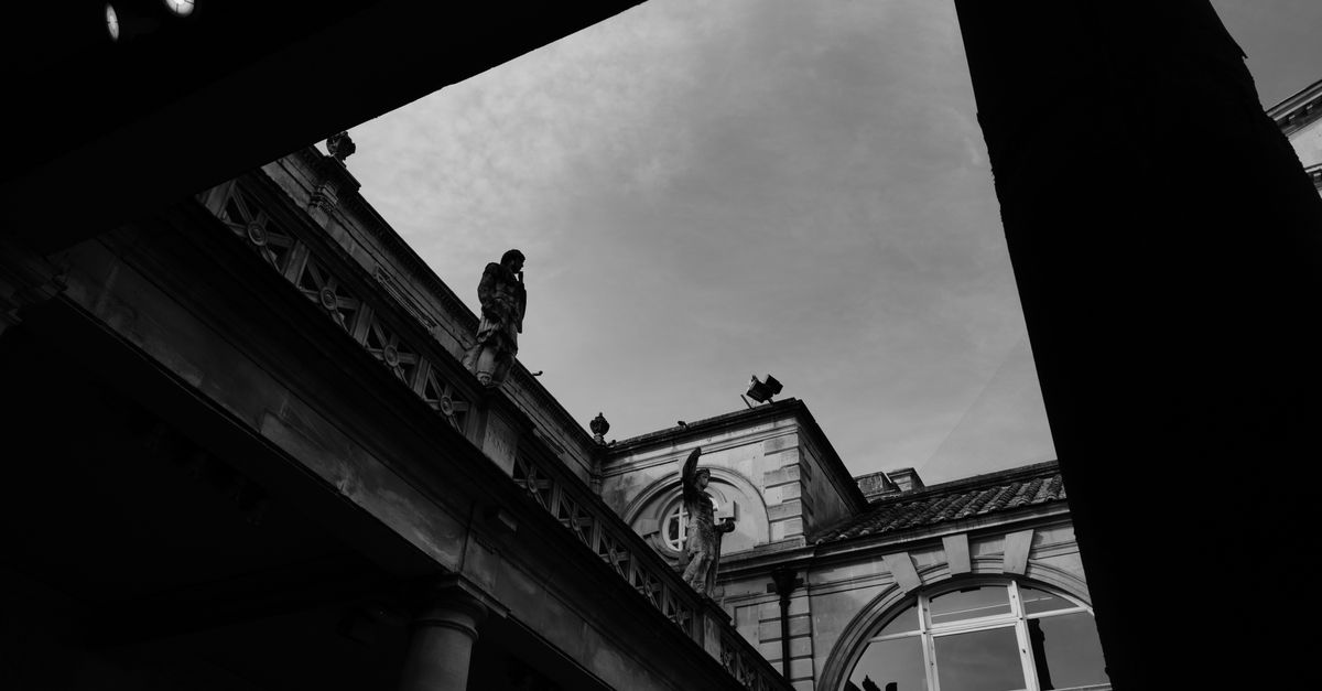 A low-angle, black-and-white view of the Roman Baths building in Bath, with classical statues silhouetted on the terrace.