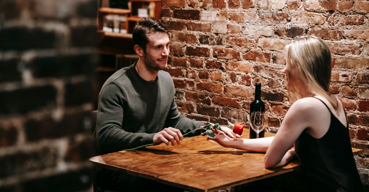 A couple sitting at a candlelit restaurant table with wine and a red rose, exposed brick behind them.